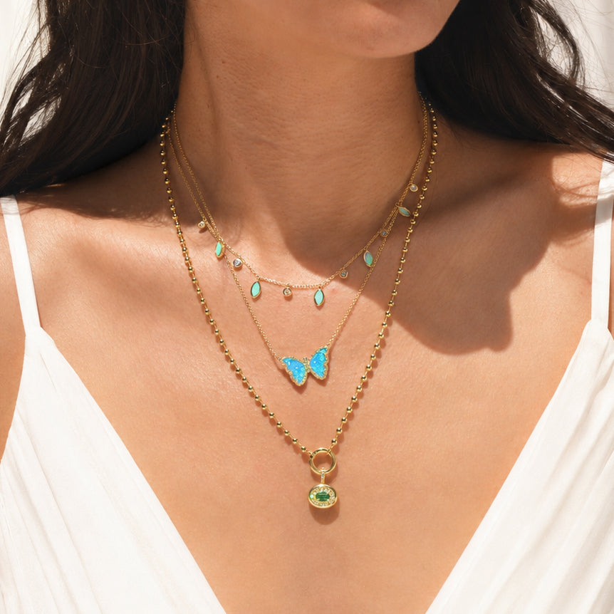 A close-up of a model wearing a layered gold necklace set. The set includes a delicate gold beaded chain with small turquoise teardrop charms, a longer chain with a light blue butterfly pendant, and a gold chain with a green gemstone pendant.