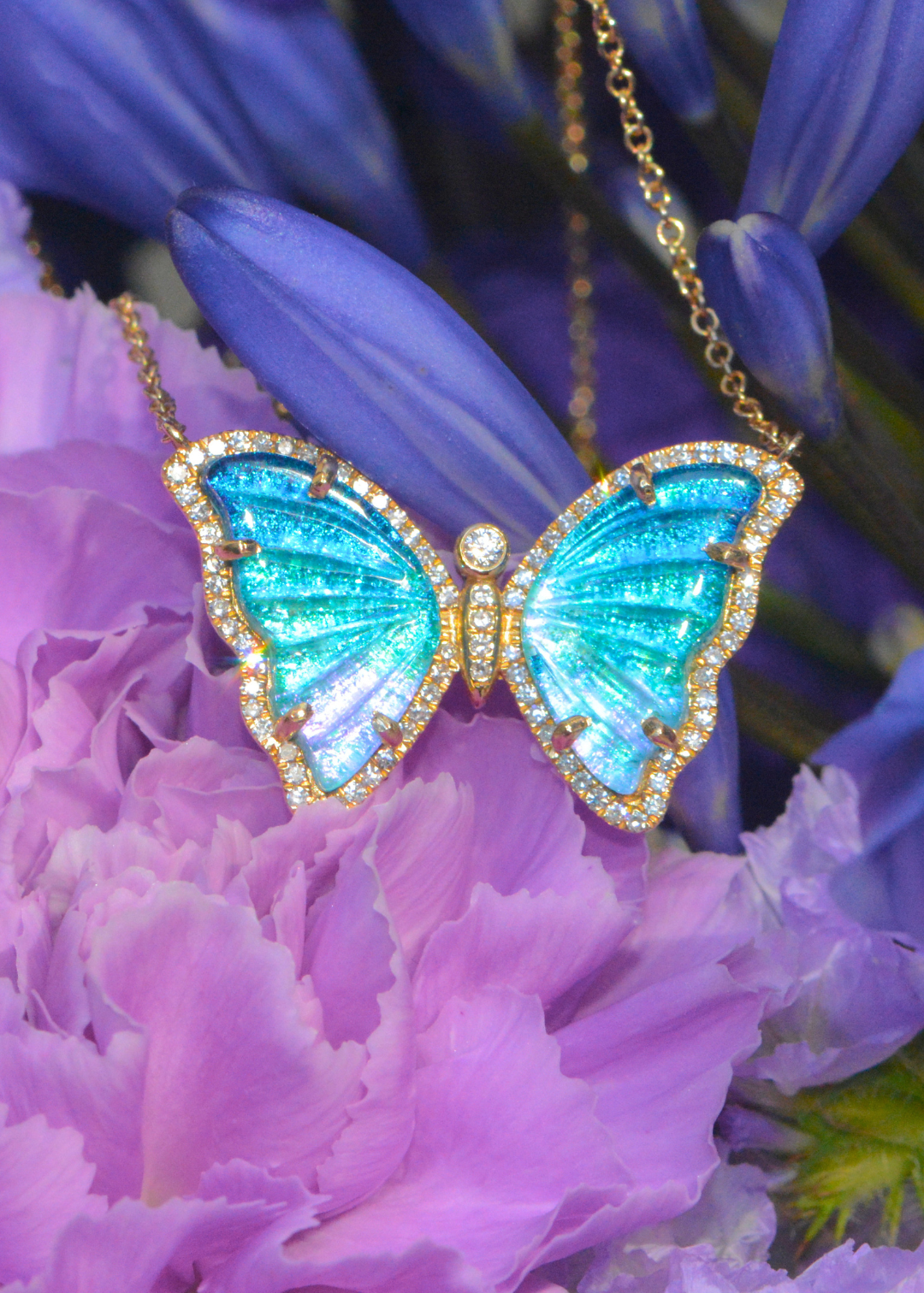 A gold diamond butterfly pendant necklace with iridescent turquoise-blue wings and small clear stones, displayed against purple flowers.